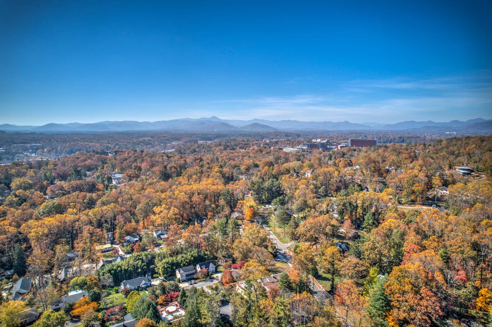 Aerial view of Asheville neighborhoods in peak fall color with the downtown skyline and Blue Ridge mountains on the horizon