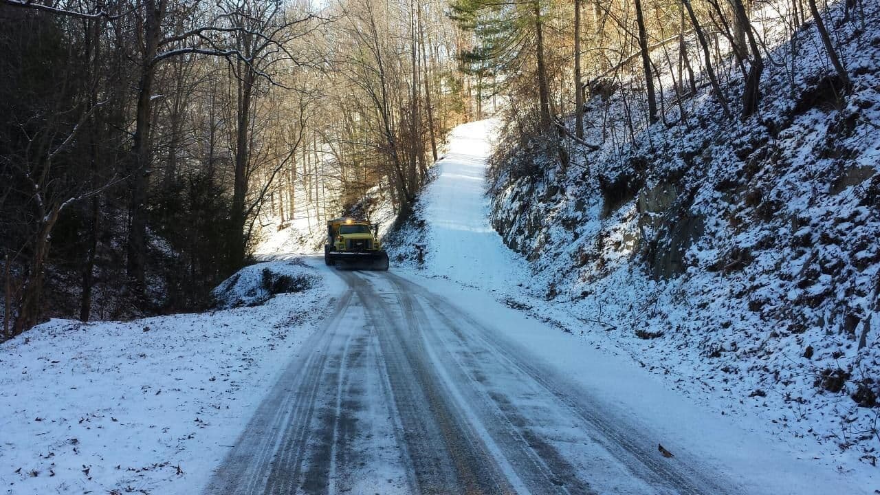 NCDOT truck plowing a narrow, snow-covered mountain road lined with bare trees
