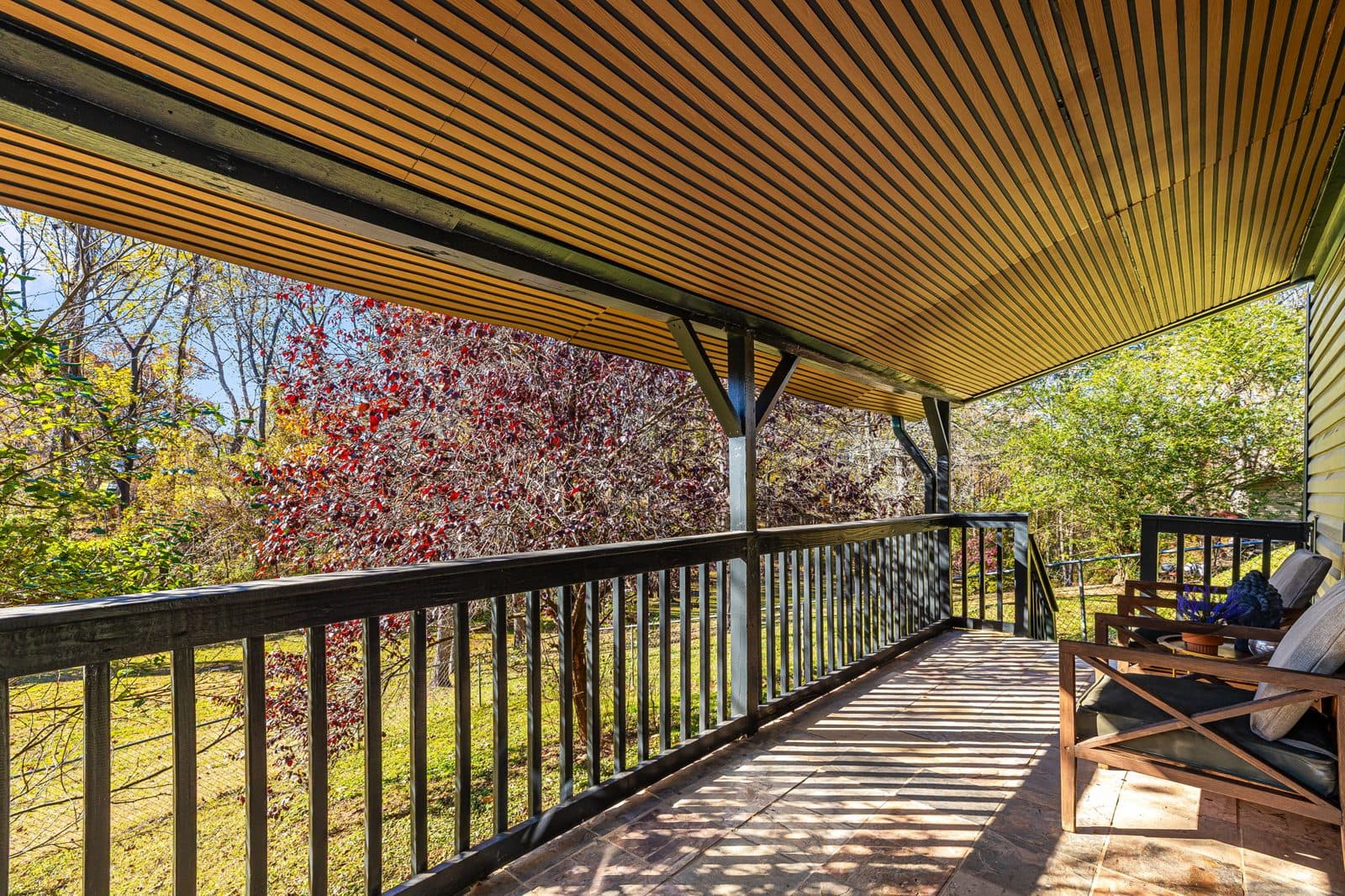 Covered wooden deck with a slatted ceiling and railing, looking out into autumn trees