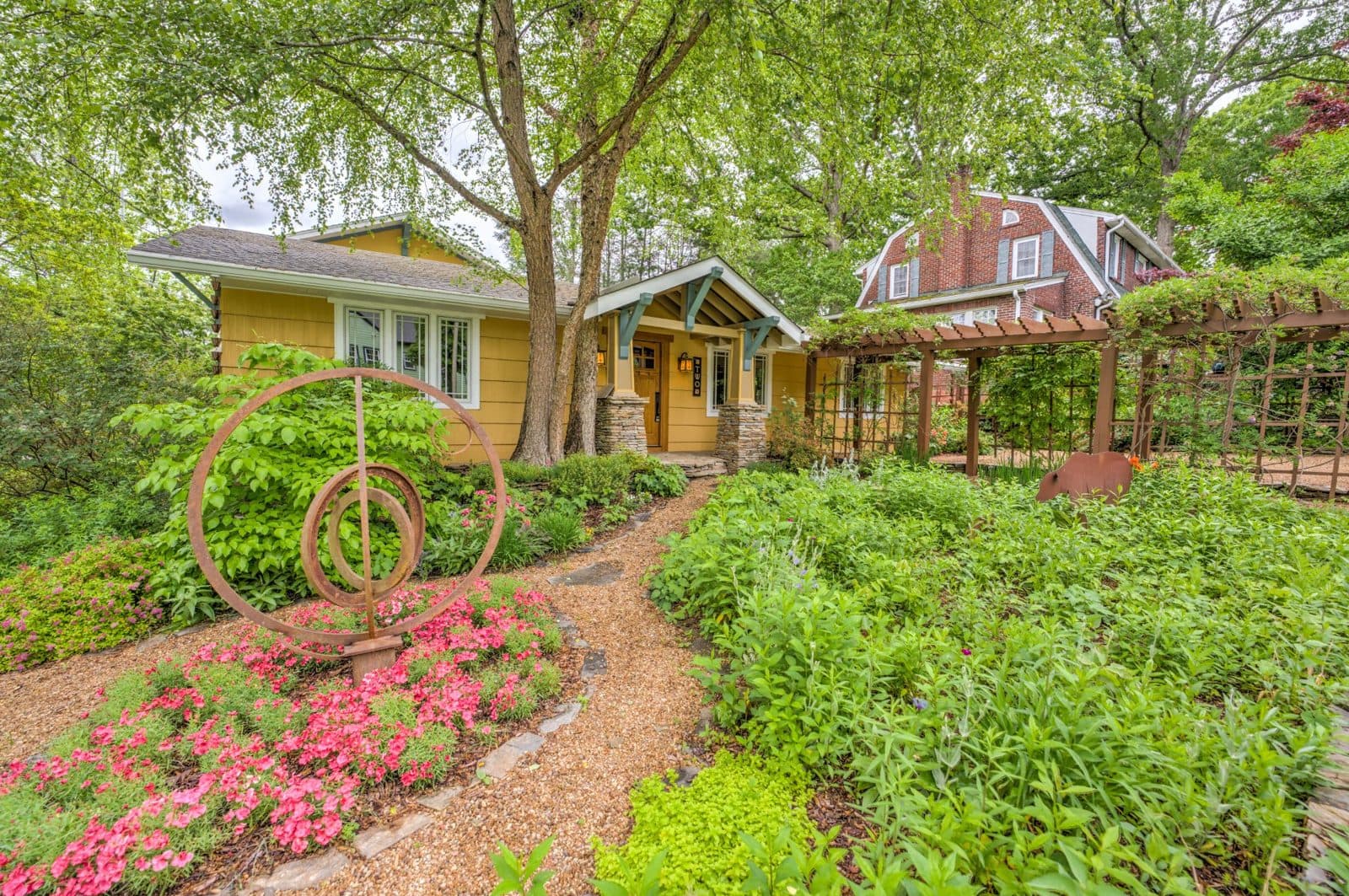 Yellow craftsman-style home behind a lush flowering garden and a wooden pergola