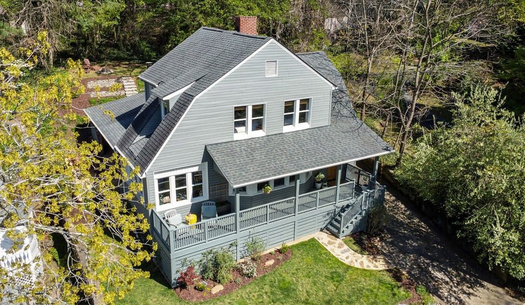 Gray two-story home with a wraparound porch on a sloped, wooded lot, with steps down to a lower yard
