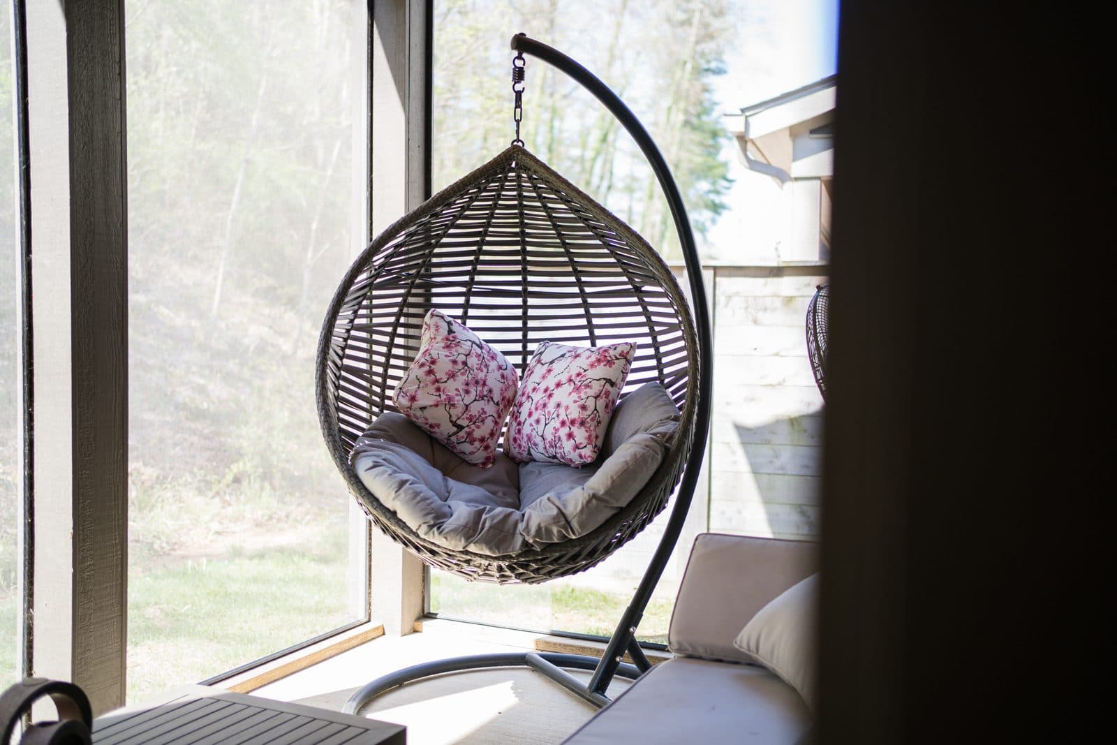 Hanging wicker egg chair with pillows beside a floor-to-ceiling window looking into the woods