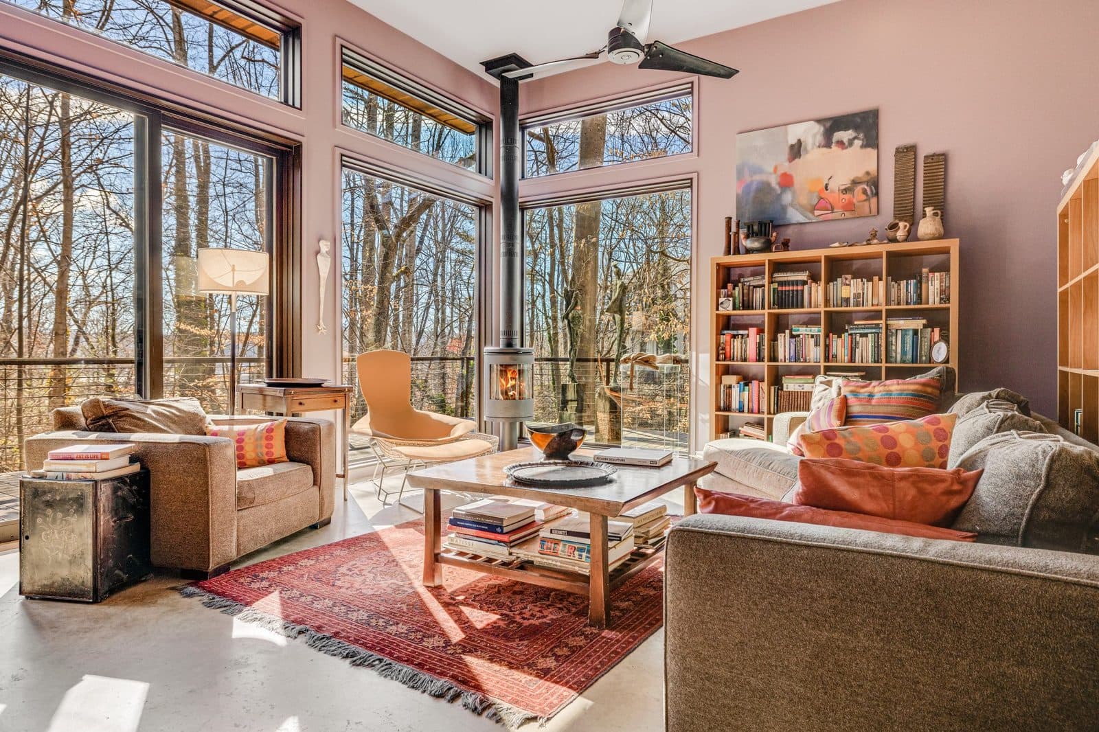 Sunroom with floor-to-ceiling windows onto bare winter trees, a wood stove, bookshelves and armchairs