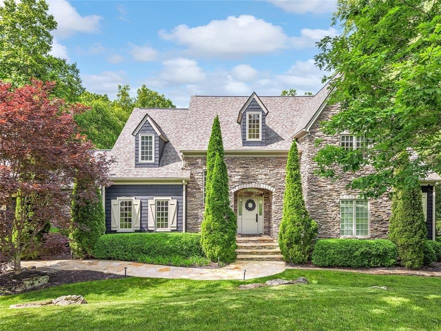 Stone and shingle home with dormers, tall evergreens and a wide green lawn