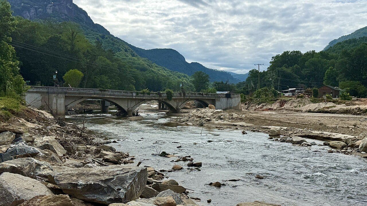 Broken remains of the Lake Lure Flowering Bridge over a rocky river channel, with mountains behind